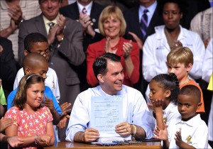 Wisconsin Governor Scott Walker presents the state budget after signing while surrounded by children from the Boys & Girls Club of Kenosha during a private ceremony at Catalyst Exhibits Sunday, July 30, in Pleasant Prairie, Wis. The budget approved by the Republican-controlled Legislature includes all Walker's priorities, including a $650 million income tax cut, expansion of private school vouchers and changes to the state's Medicaid and food stamp programs. (AP Photo/Kenosha News, Brian Passino)