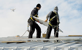 A drone flies overhead as Marcus Lisocki (left) and Kyle Cruver, both employees of West Bend-based Steiner Electric, install solar panels on the roof of the Ozaukee County Transit Services building in Port Washington. Drone pilots may soon find that failure to follow the changing rules can have severe consequences. (Staff Photo by Kevin Harnack) 