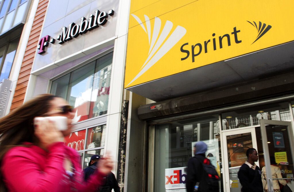 FILE - In this April 27, 2010 file photo, a woman using a cell phone walks past T-Mobile and Sprint stores in New York. Published reports say a group of state attorneys general are planning a lawsuit to block a $26.5 billion merger of wireless carriers T-Mobile and Sprint. It’s an unusual step ahead of a decision by federal antitrust authorities. (AP Photo/Mark Lennihan, File)