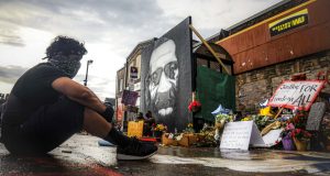 After a new mural, center, of George Floyd is added to a growing memorial of tributes, Trevor Rodriquez sits alone at the spot where Floyd died while in police custody, Tuesday June 2, 2020, in Minneapolis, Minn. "I have been out every single night protesting peacefully, just trying to support everything," said Rodriquez. "I didn't want to come here just on a rush, so I had to just take a moment to pay my respect." (AP Photo/Bebeto Matthews)