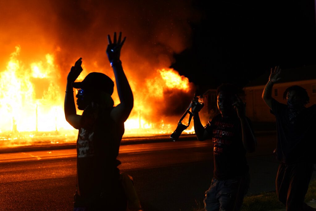 FILE - In this Aug. 24, 2020, file photo, protesters walk past police with their arms up, in Kenosha, Wis., as a building burns in the background. Protests have erupted following the police shooting of Jacob Blake a day earlier. Kenosha police say 55 people are facing charges related to violent demonstrations after the police shooting of Jacob Blake last summer. (AP Photo/David Goldman, File)